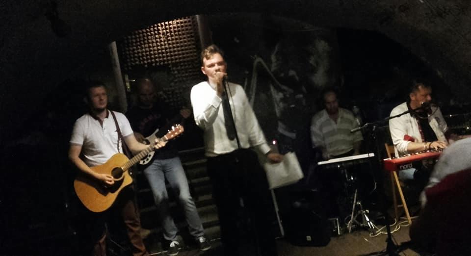 Zespół grający koncert w piwnicy z surowymi ścianami, wokalista w białej koszuli i czarnym krawacie śpiewa do mikrofonu, gitarzysta z gitarą akustyczną, klawiszowcy grają na keyboardach.