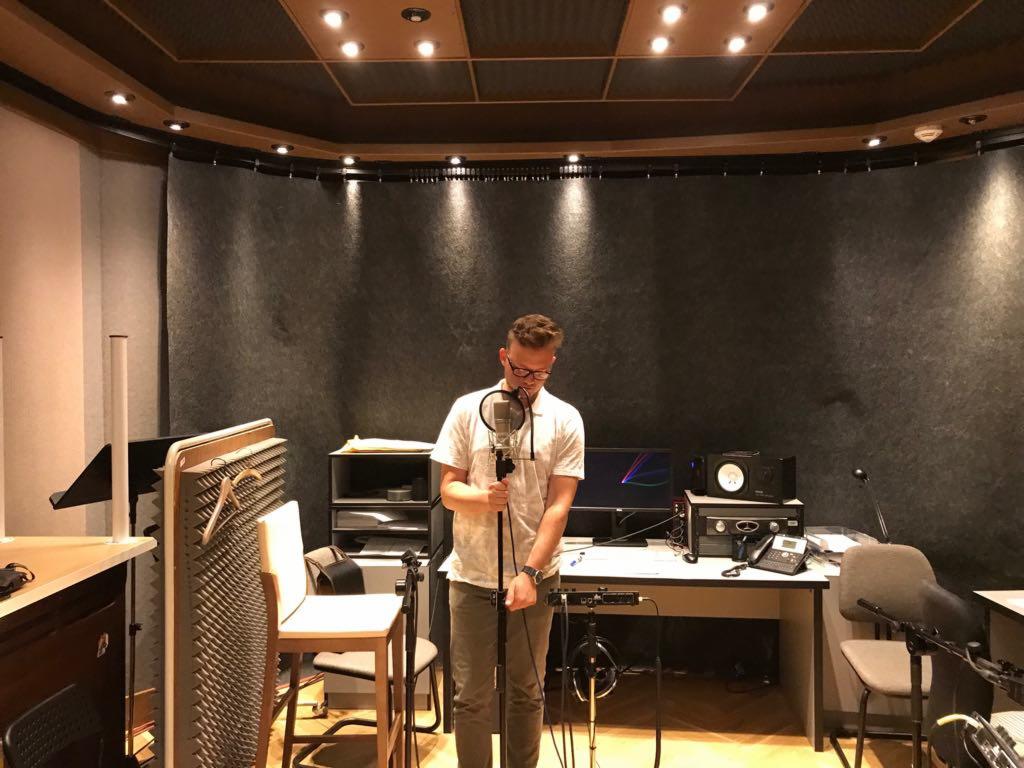 Młody mężczyzna w okularach przygotowuje mikrofon w profesjonalnym studiu nagraniowym z panelami akustycznymi na ścianach i suficie, biurkiem z monitorem i sprzętem audio w tle.