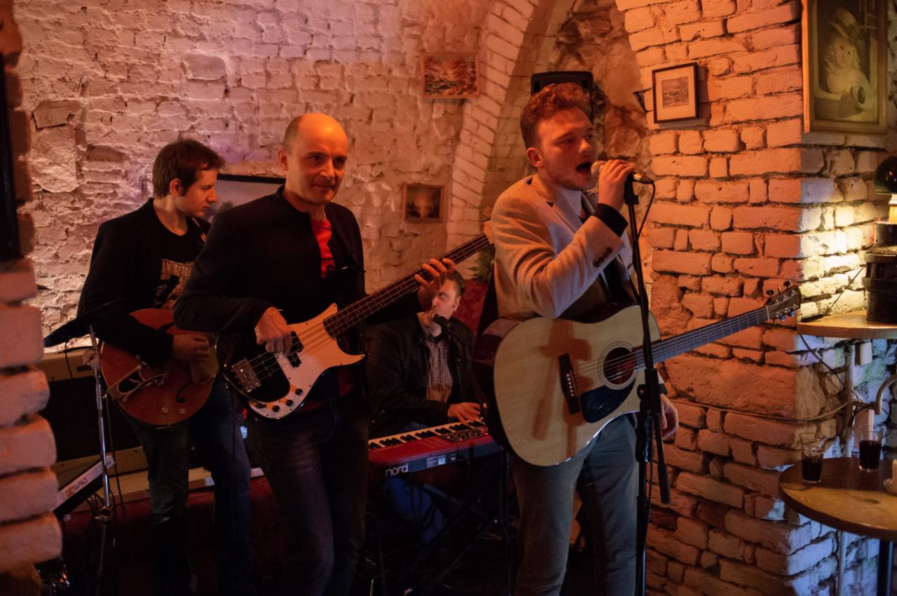 Zespół muzyczny grający koncert w ceglanym wnętrzu, wokalista śpiewa do mikrofonu, gitarzysta basowy i gitarzysta grają na instrumentach, klawiszowiec w tle.