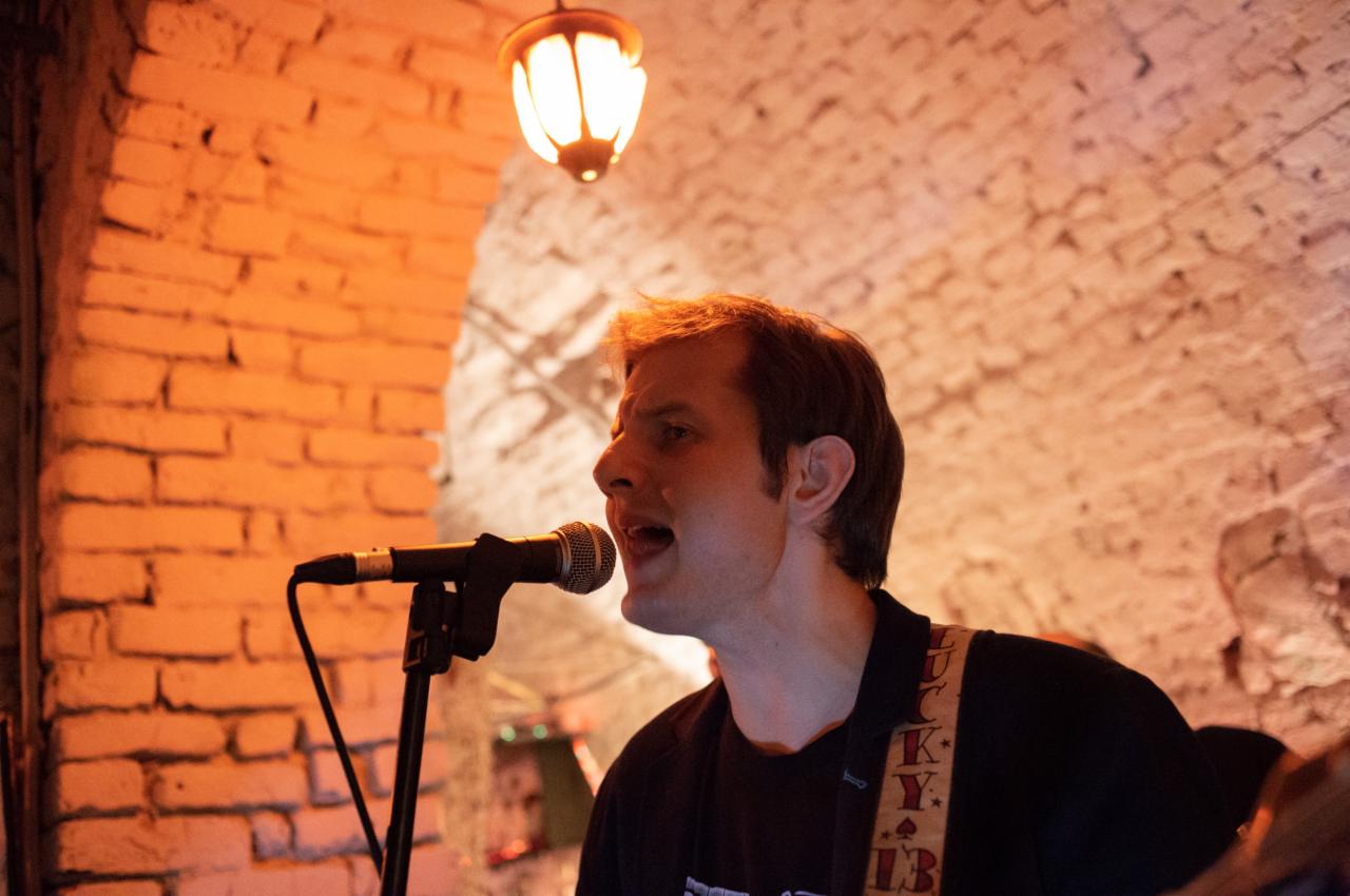 Wokalista śpiewa do mikrofonu na tle ceglanej ściany i lampy, pasek gitary z motywem kart.