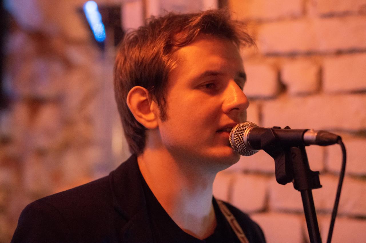 Portret młodego mężczyzny śpiewającego do mikrofonu na tle ceglanej ściany oświetlonej ciepłym światłem.