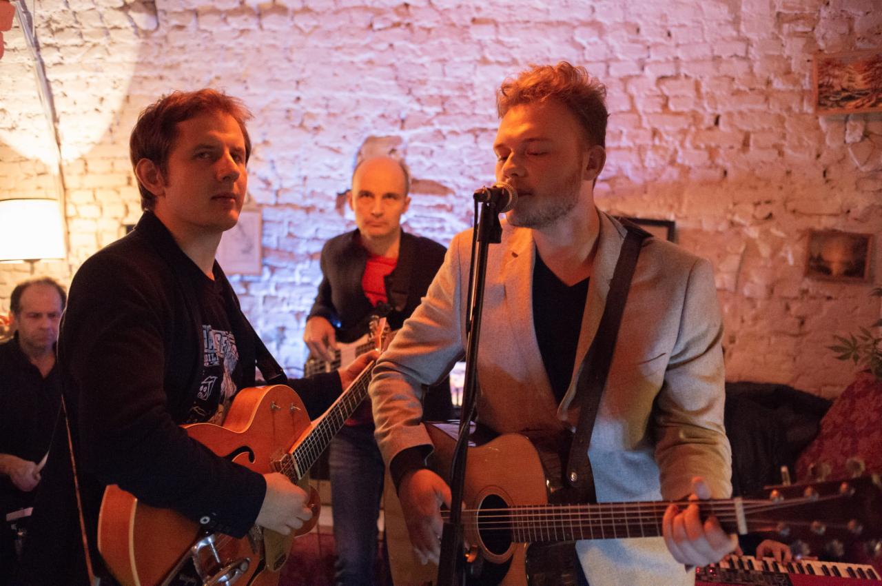 Zespół grający muzykę na żywo w klubie z ceglaną ścianą w tle, wokalista śpiewa do mikrofonu, gitarzysta gra na gitarze typu hollow body, basista w tle.