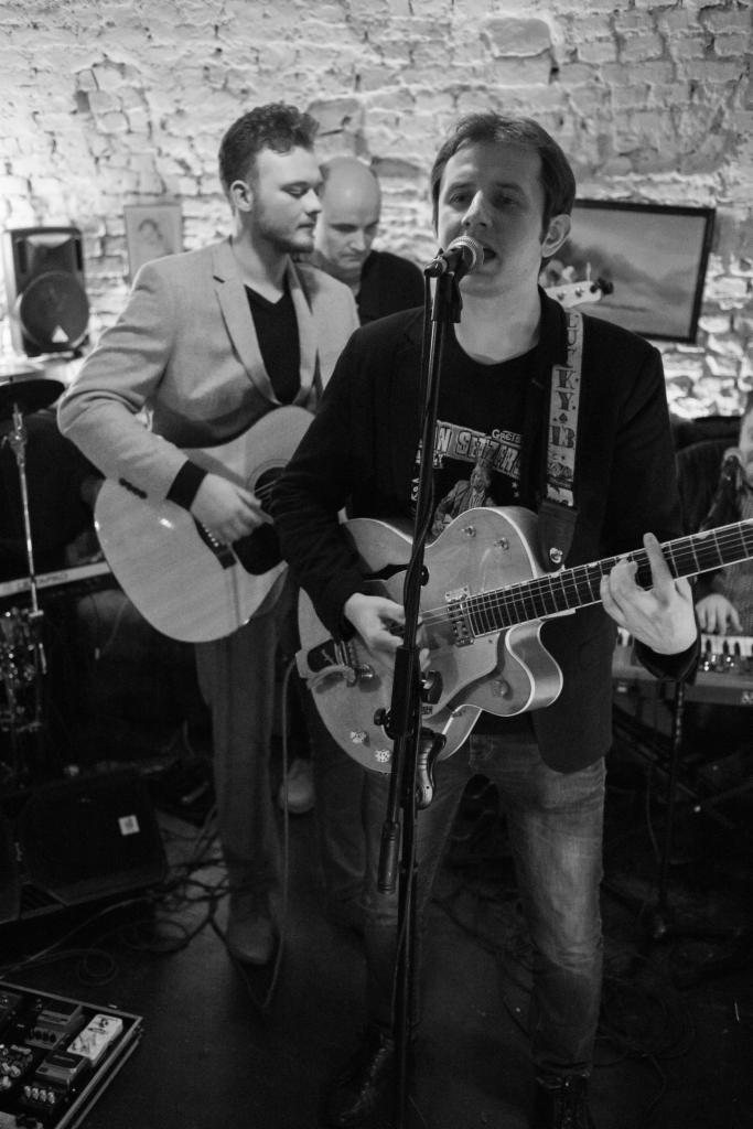 Czarno-białe zdjęcie zespołu muzycznego podczas występu na żywo w lokalu z ceglaną ścianą, wokalista śpiewa do mikrofonu grając na gitarze elektrycznej, gitarzysta akompaniuje na gitarze...