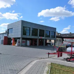 Budynek centrum rolniczego z szyldem reklamującym mleczarstwo, drobiarstwo, maszyny rolnicze, hale namiotowe i fotowoltaikę, na pierwszym planie maszyna rolnicza.