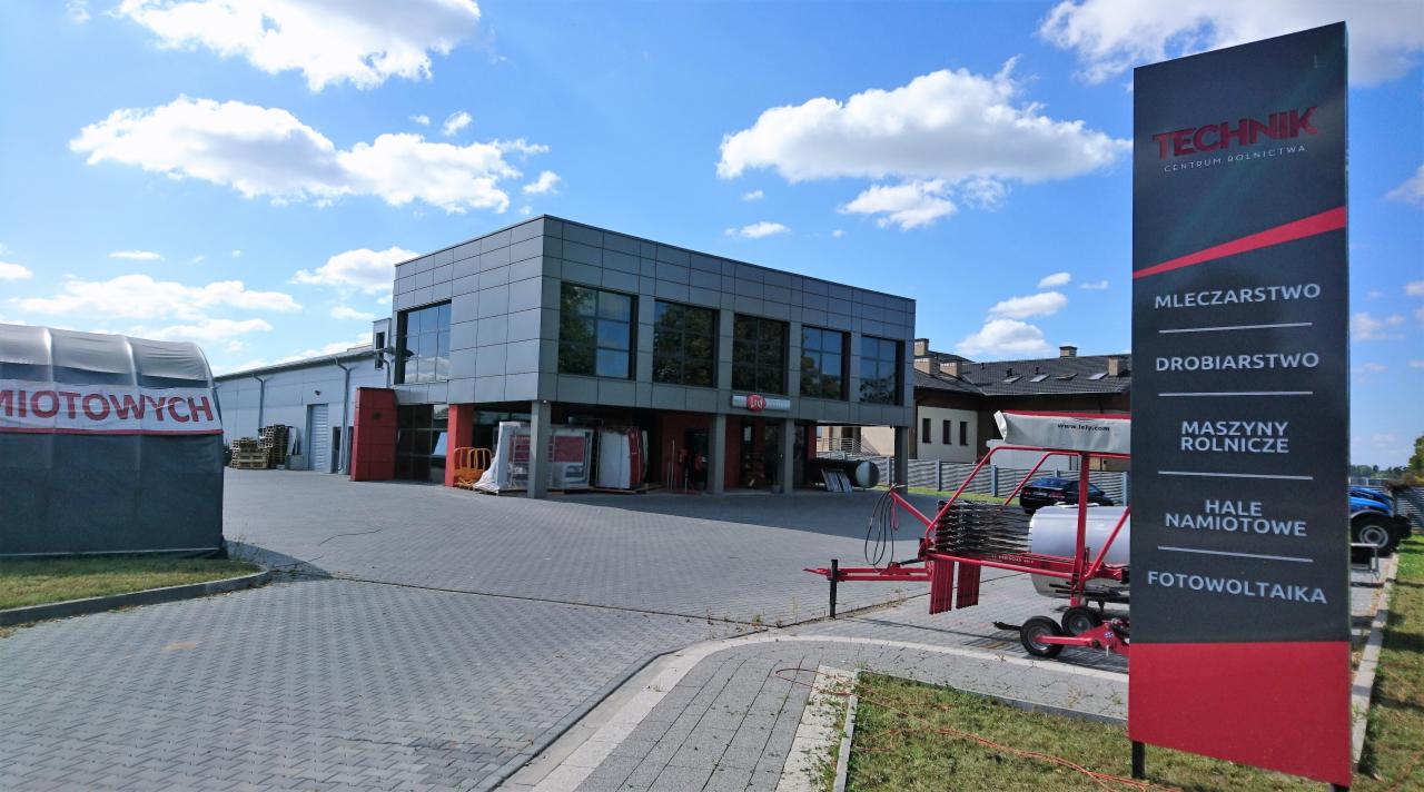 Budynek centrum rolniczego z szyldem reklamującym mleczarstwo, drobiarstwo, maszyny rolnicze, hale namiotowe i fotowoltaikę, na pierwszym planie maszyna rolnicza.