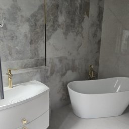Dolecińska Projekt DARIA DOLECIŃSKA - Nowoczesna łazienka z wolnostojącą wanną, marmurowymi płytkami i złotą armaturą. Umywalka z szafką w kształcie łuku. Widok na okno.