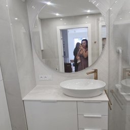 Dolecińska Projekt DARIA DOLECIŃSKA - Nowoczesna łazienka z okrągłym lustrem i białą szafką pod umywalkę. Złota bateria i szare płytki. W lustrze odbija się osoba robiąca zdjęcie.