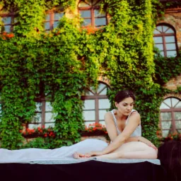 Masaż pleców wykonywany przez masażystkę na tle ceglanego budynku porośniętego bluszczem i z czerwonymi kwiatami w oknach.