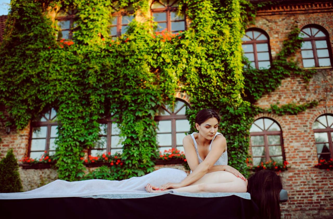 Masaż pleców wykonywany przez masażystkę na tle ceglanego budynku porośniętego bluszczem i z czerwonymi kwiatami w oknach.