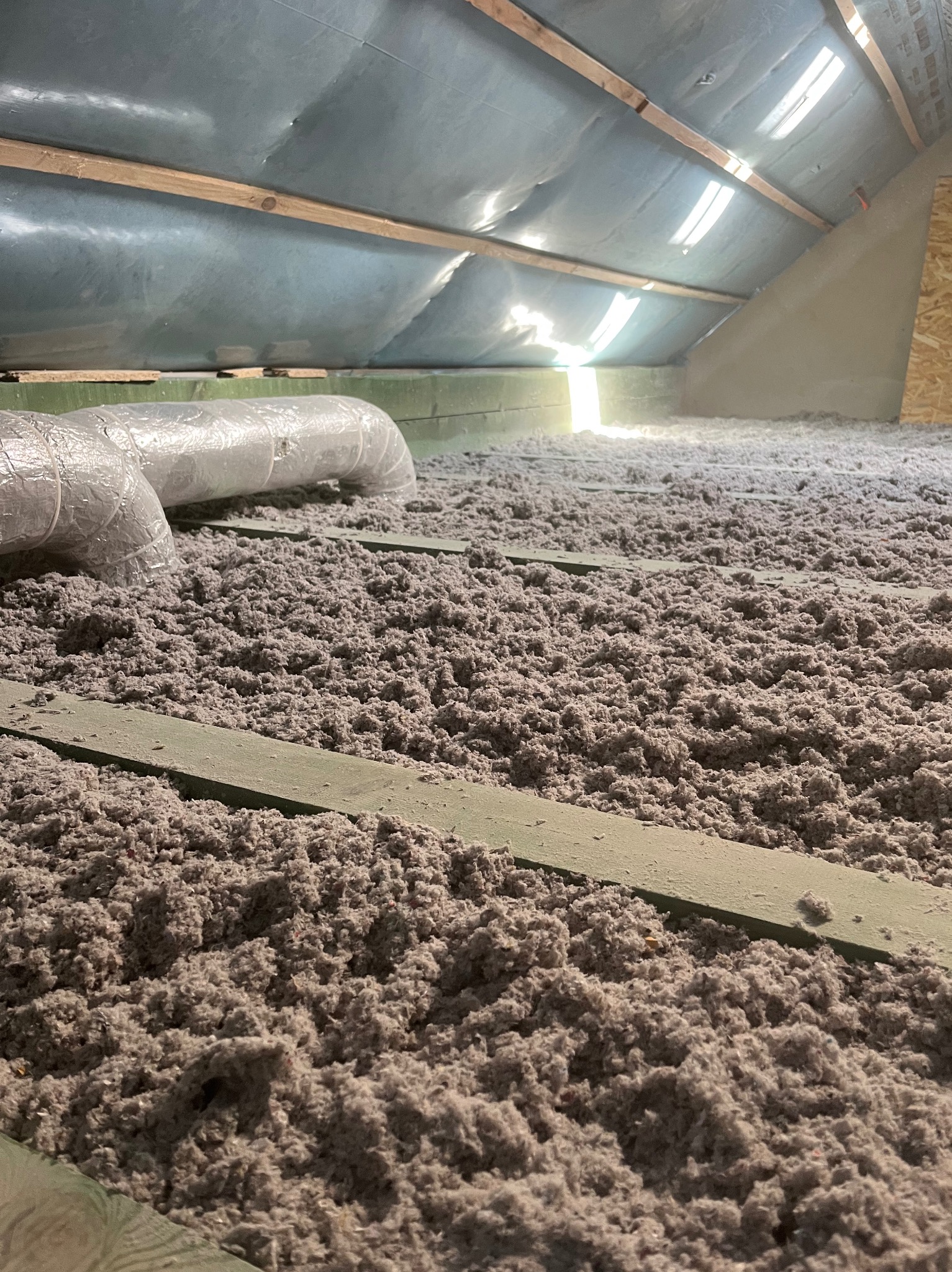 Ocieplenie poddasza wełną celulozową z widoczną instalacją wentylacyjną i drewnianą konstrukcją dachu. Ujęcie z niskiego kąta.