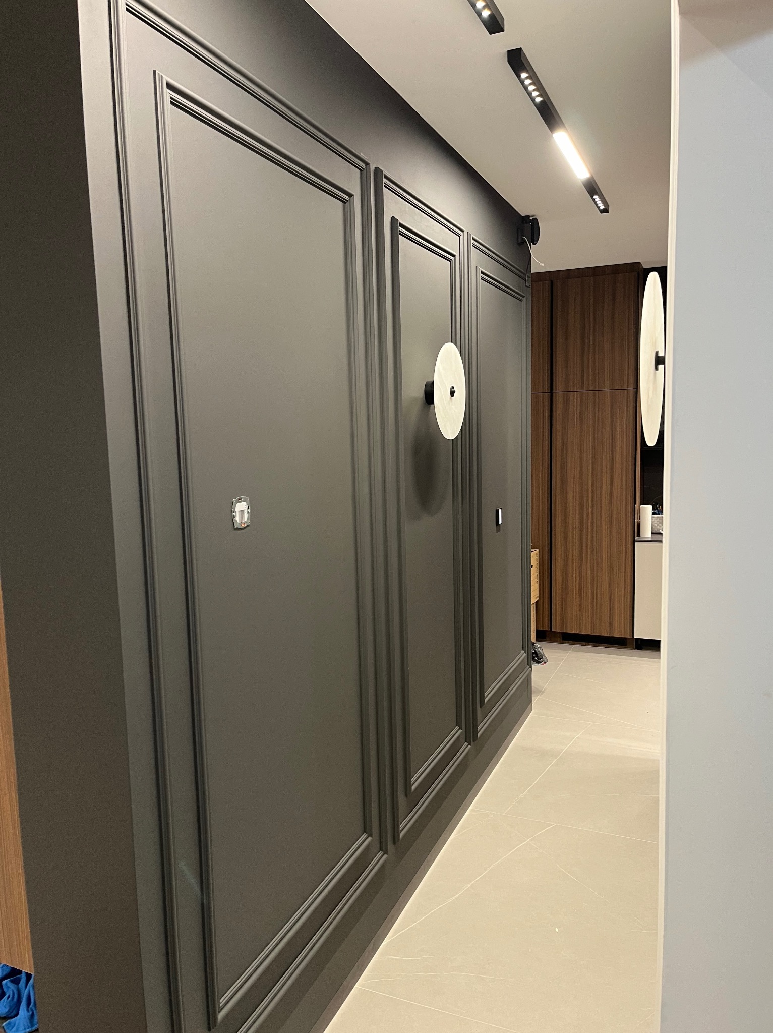 Ciemnoszara, panelowa ściana z dekoracyjnymi listwami i okrągłymi lampami ściennymi. Widoczny fragment podłogi z jasnych płytek i fragment zabudowy kuchennej w tle.