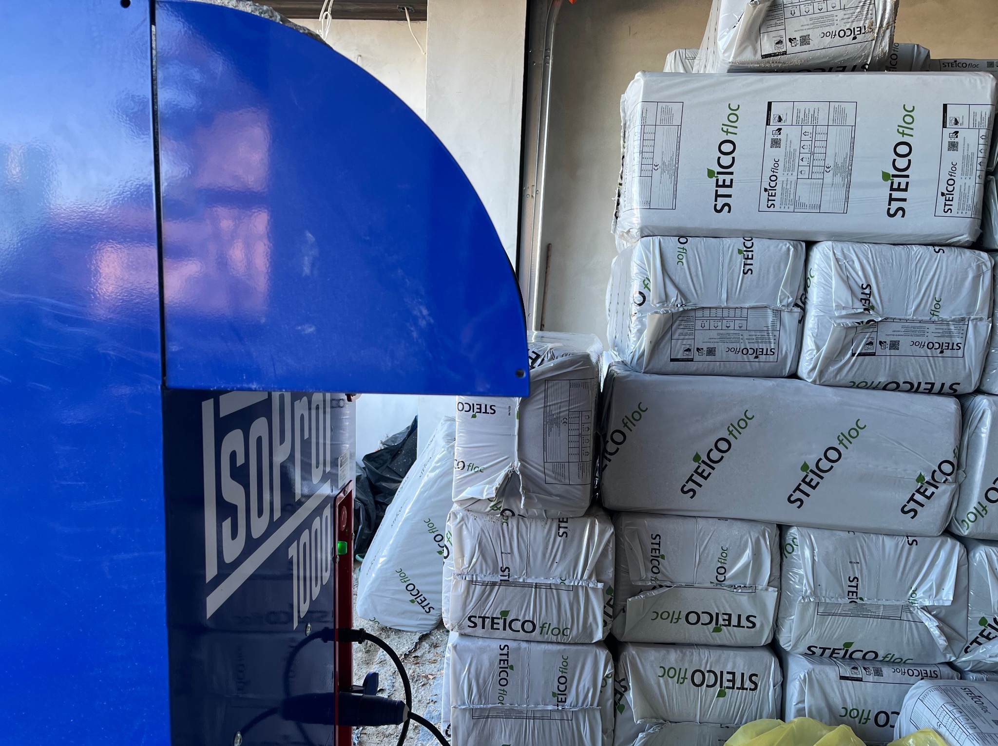 Stos worków izolacyjnych Steico floc obok niebieskiego urządzenia ISOP Pearl 1000, w surowym wnętrzu, prawdopodobnie przed montażem.