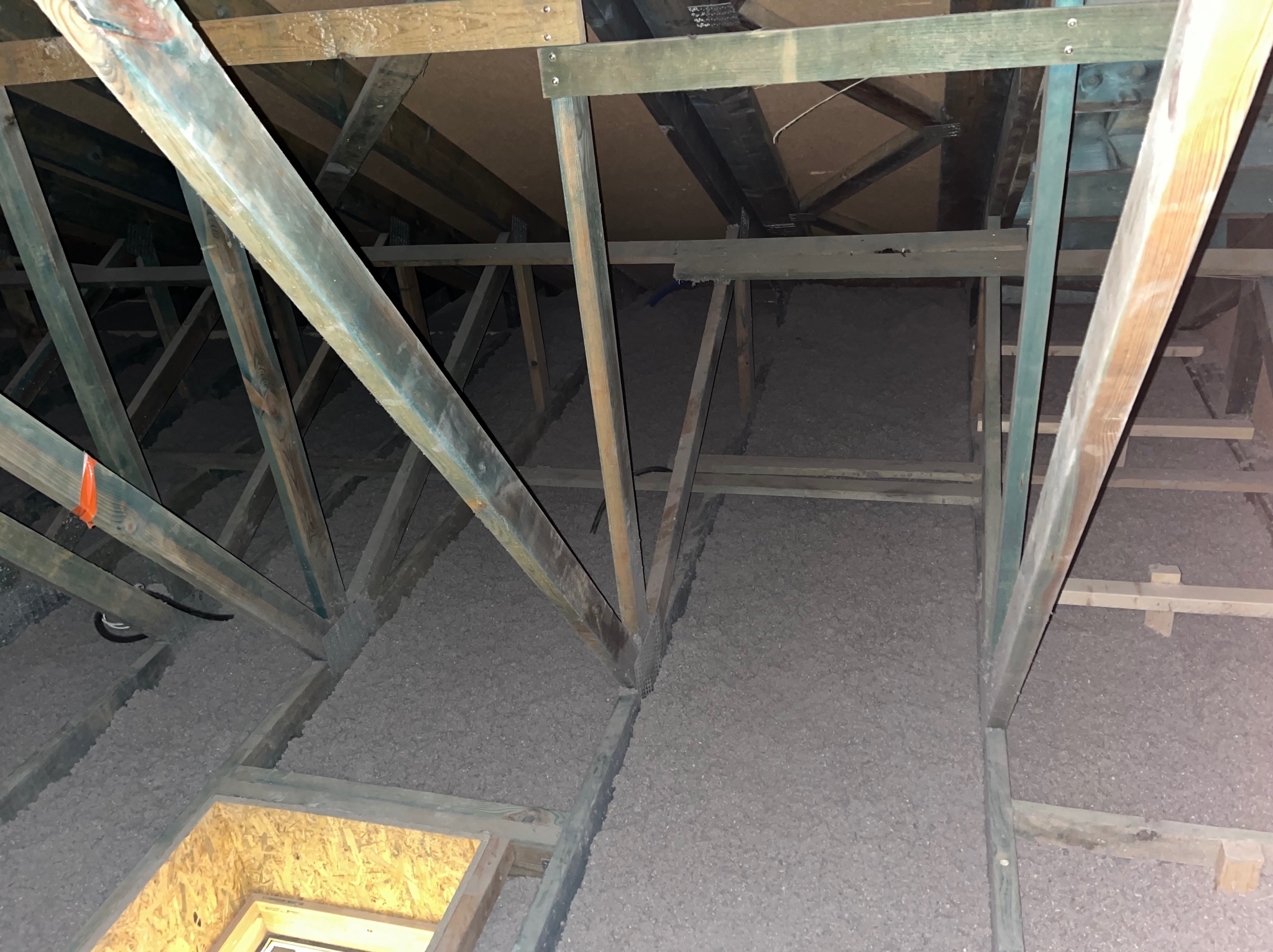 Ocieplenie poddasza wełną mineralną, widoczne drewniane elementy konstrukcyjne dachu i warstwa izolacji termicznej. Szara wełna wypełnia przestrzeń między belkami.