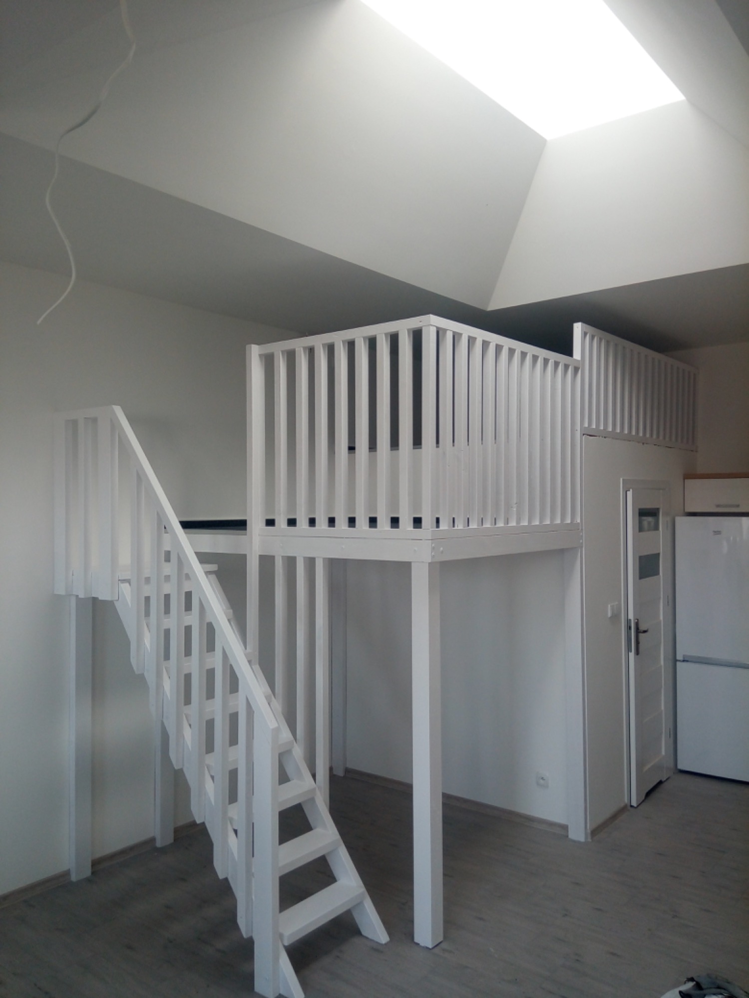 Biała antresola z drewnianymi schodami w jasnym pokoju z oknem dachowym, widoczna również biała lodówka i drzwi.