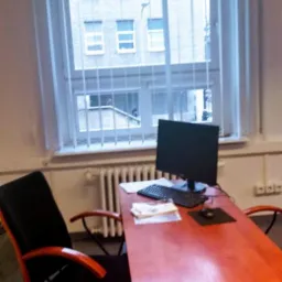 Biuro z pomarańczowym biurkiem, krzesłem, komputerem i dokumentami przed oknem z żaluzjami pionowymi.