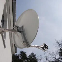 Satelitarna antena zamontowana na ścianie budynku, widok z dołu na tle błękitnego nieba i drzew bez liści.