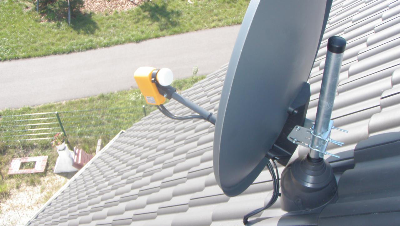 Szara antena satelitarna zamontowana na brązowym dachu z dachówki ceramicznej, widok z góry na okolicę z zieloną trawą i drogą.