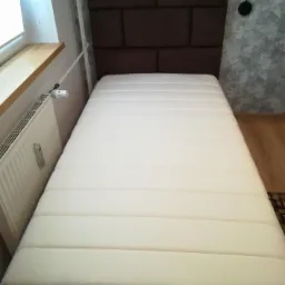 Łóżko jednoosobowe z brązowym, panelowym wezgłowiem, ustawione przy ścianie z szarą tapetą, widok z przodu.