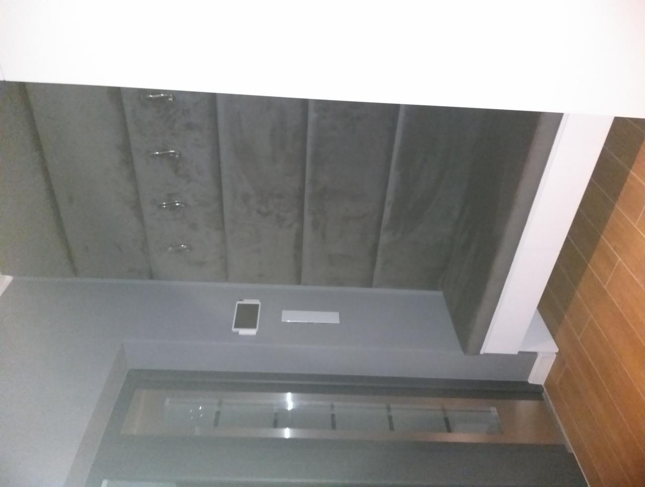 Szara, tapicerowana ściana z podłużnymi panelami i metalowymi guzikami, widoczna nad drzwiami w szarej ramie i fragmentem drewnianej podłogi.