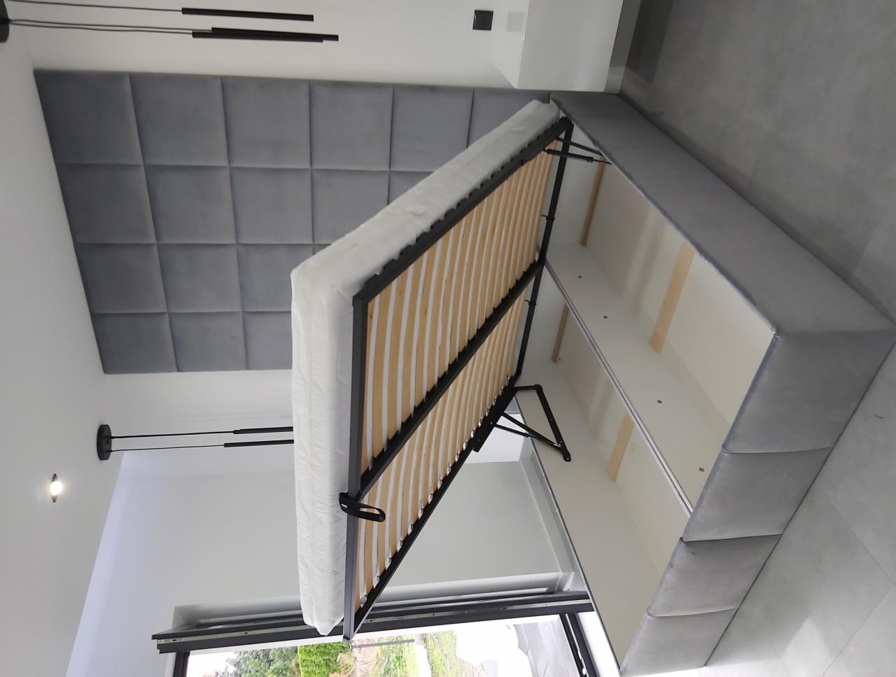 Łóżko z podnoszonym stelażem i pojemnikiem na pościel, obite szarą tkaniną, widoczne od strony stelaża i wnętrza pojemnika, z szarą tapicerowaną ścianą w tle.