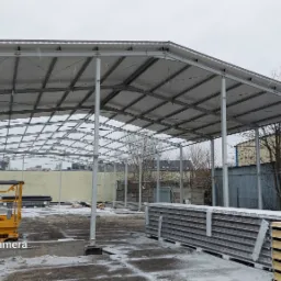 Konstrukcja stalowa zadaszenia w trakcie montażu, widoczny podnośnik Haulotte Ramirent i przygotowane płyty warstwowe na ziemi pokrytej śniegiem.