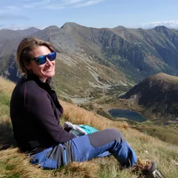 Uśmiechnięta kobieta w okularach przeciwsłonecznych siedzi na trawiastym zboczu góry z widokiem na pasmo górskie i jezioro w dolinie.