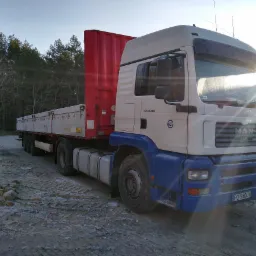 Ciężarówka MAN z czerwoną naczepą kontenerową i platformą, zaparkowana na placu z kamieniami, w tle las i promienie wschodzącego słońca.