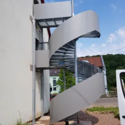 Zewnętrzne schody spiralne z metalu, prowadzące do wejścia w budynku, z widoczną konstrukcją nośną i szarymi panelami bocznymi, zamontowane przy białej ścianie budynku na zewnątrz, z fragmentem...