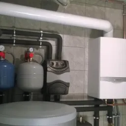 Kompaktowy kocioł grzewczy Vaillant zamontowany na kafelkowanej ścianie, widoczne rury miedziane, zbiornik wyr&oacute;wnawczy w kolorze niebieskim i szarym, oraz szary akumulator ciepła.