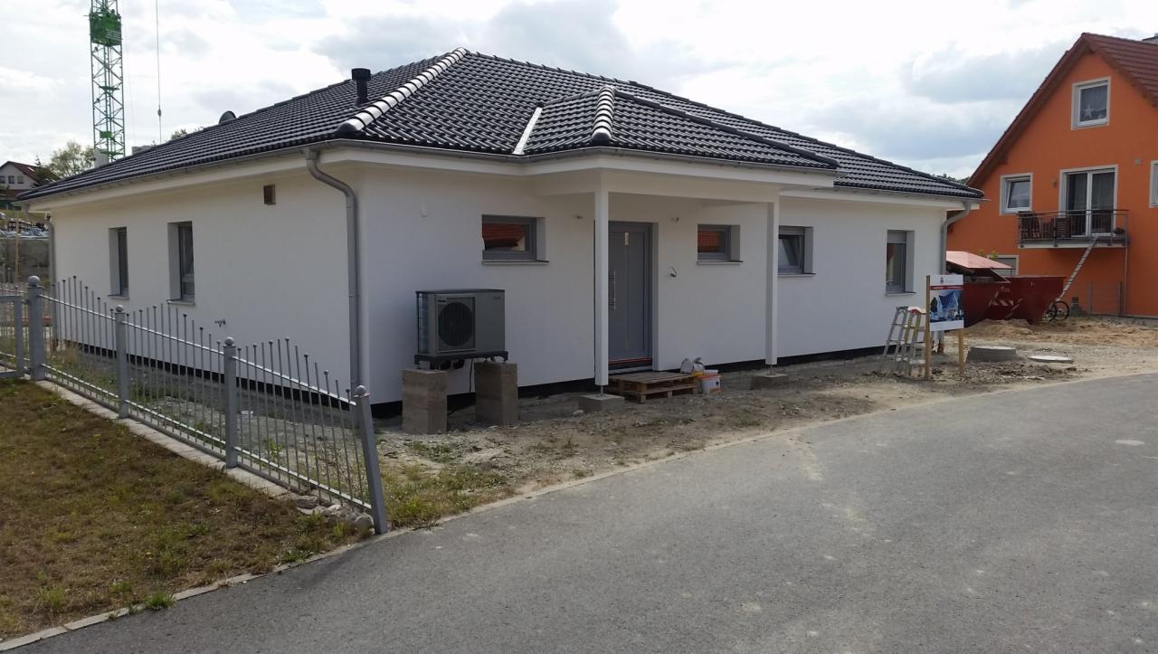 Nowo wybudowany dom jednorodzinny z zamontowaną pompą ciepła Nibe na betonowych bloczkach, widoczną z zewnątrz obok wejścia. Teren wokół domu częściowo zagospodarowany, widoczny szary chodnik...