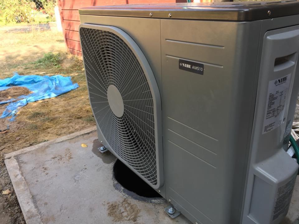 Szara pompa ciepła Nibe AMS10 zainstalowana na betonowej podstawie z widocznym otworem, na zewnątrz budynku.