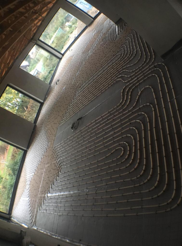 Rury ogrzewania podłogowego ułożone w pętle na szarej wylewce, widok z boku, duże okna w tle, surowy stan wykończenia wnętrza.