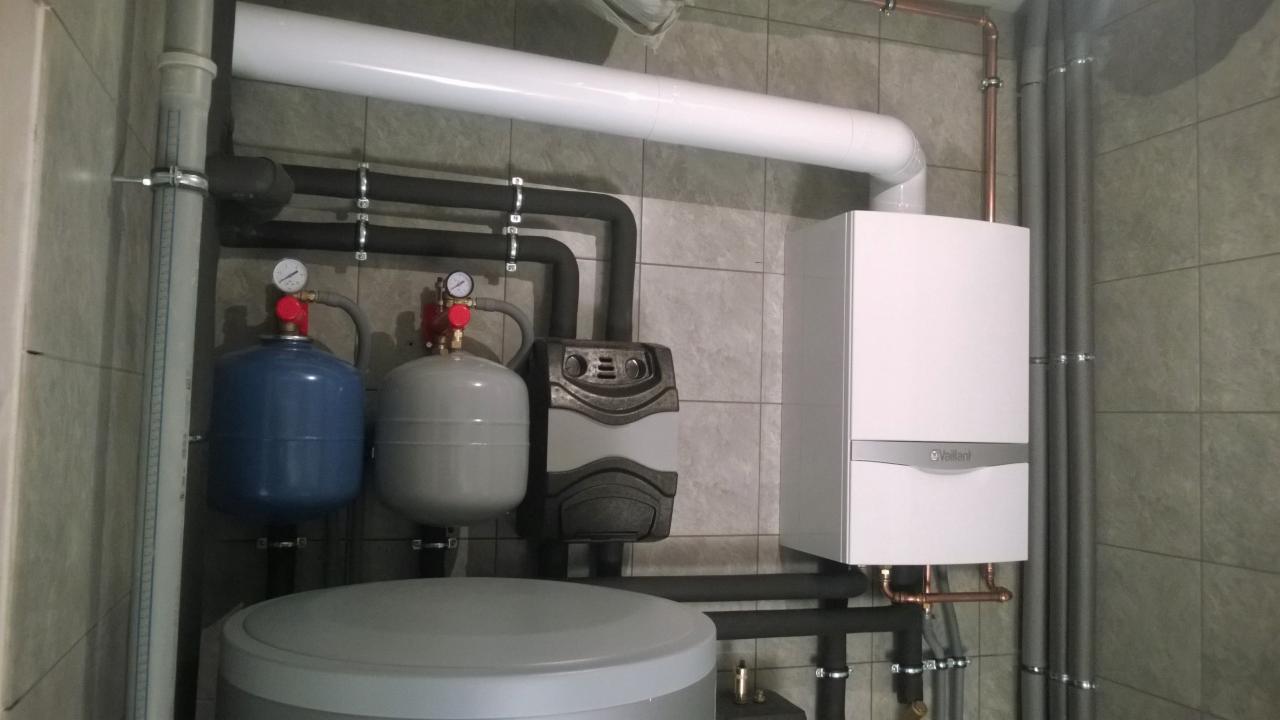 Kompaktowy kocioł grzewczy Vaillant zamontowany na kafelkowanej ścianie, widoczne rury miedziane, zbiornik wyrównawczy w kolorze niebieskim i szarym, oraz szary akumulator ciepła.