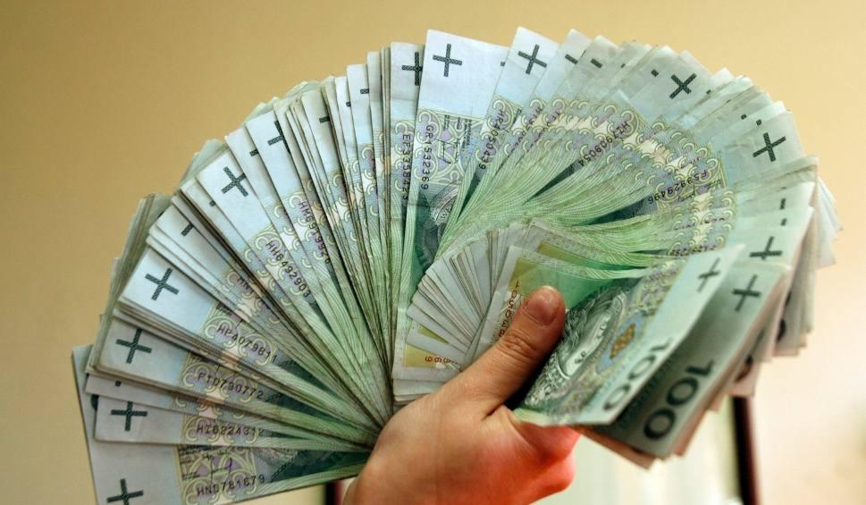 Dłoń trzymająca wachlarz banknotów 100 zł z widocznymi numerami seryjnymi, na rozmytym, beżowym tle.