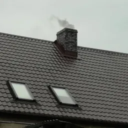 Ciemnobrązowy dach z dachówki ceramicznej z dwoma oknami dachowymi i ceglanym kominem, z którego wydobywa się dym na tle pochmurnego nieba.
