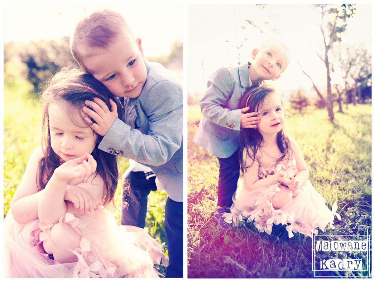 Portret dwójki dzieci w plenerze: chłopiec w jasnoszarym żakiecie obejmuje dziewczynkę w różowej sukience z falbanami, na trawie, w tle rozmyte drzewa i słońce.