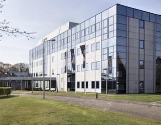 Elewacja nowoczesnego, wielopiętrowego biurowca z licznymi oknami odbijającymi niebo, przed budynkiem zadbany trawnik i chodnik.