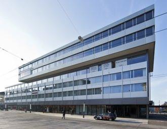 Nowoczesny, wielopiętrowy budynek biurowy z licznymi oknami i metalową elewacją, z widocznym fragmentem ulicy i przechodniami. Charakterystyczny element architektoniczny - wysunięta bryła górnych...