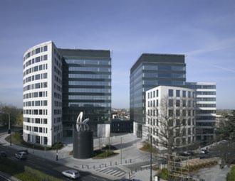 Nowoczesny kompleks biurowy z trzema budynkami o zróżnicowanej architekturze: zaokrąglony, prostopadłościan i wieża, połączone placem z czarną, cylindryczną rzeźbą.