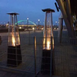 Dwa stojące grzejniki gazowe piramidalne z widocznym płomieniem, ustawione na zewnątrz na tle wieczornego miasta z oświetlonym mostem.