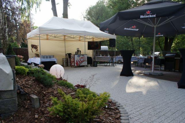 Uroczystość plenerowa: biały namiot z wyposażeniem barowym, stoliki koktajlowe pod parasolami Heineken, oświetlona kula dekoracyjna i zadbana roślinność.