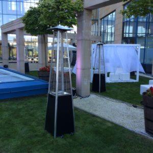 Dwa stalowe promienniki ciepła w kształcie piramidy na trawniku obok białego namiotu imprezowego i basenu, na tle nowoczesnego budynku z kolumnami.