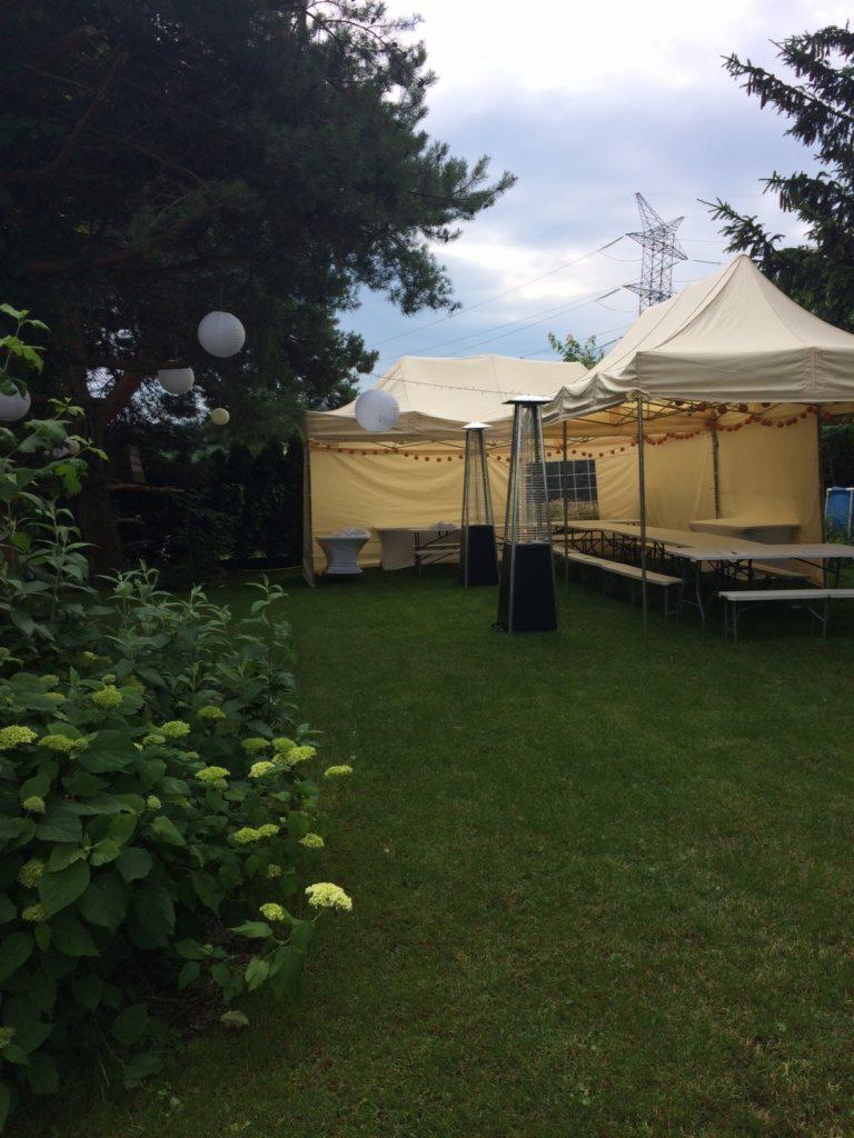 Ustawione namioty imprezowe z oświetleniem i ogrzewaniem na trawniku, widoczne wiszące lampiony i słupy wysokiego napięcia w tle.