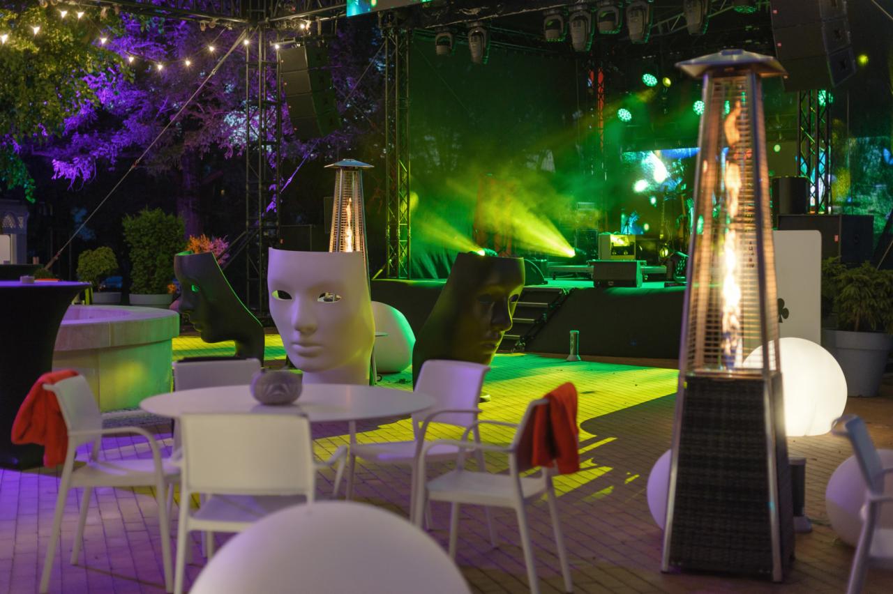 Scena plenerowa z oświetleniem, stoły z krzesłami, dekoracyjne maski i promiennik ciepła, przygotowana na imprezę.