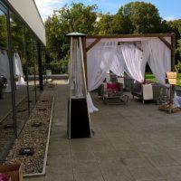 Zadaszona przestrzeń wypoczynkowa z białymi zasłonami, drewnianą konstrukcją i grzejnikiem gazowym na tarasie z widokiem na zieleń.