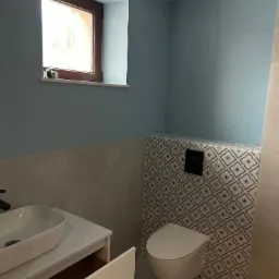 Wykończona łazienka w odcieniach szarości i błękitu z wiszącą toaletą na tle wzorzystych płytek i umywalką na szafce z otwartą szufladą.