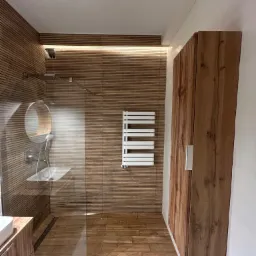 Nowoczesna łazienka z kabiną prysznicową typu walk-in, wykończona płytkami imitującymi drewno, z grzejnikiem drabinkowym i szafką z frontami drewnopodobnymi.