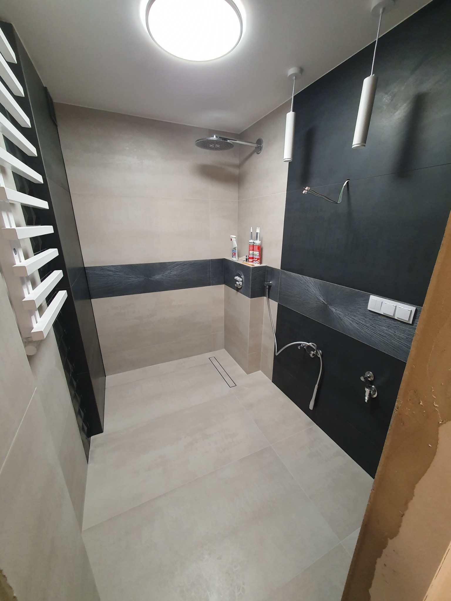 Nowoczesna kabina prysznicowa z czarno-beżowymi płytkami, deszczownicą, grzejnikiem drabinkowym i wiszącymi lampami. Widoczne niedokończone prace wykończeniowe przy framudze drzwi.