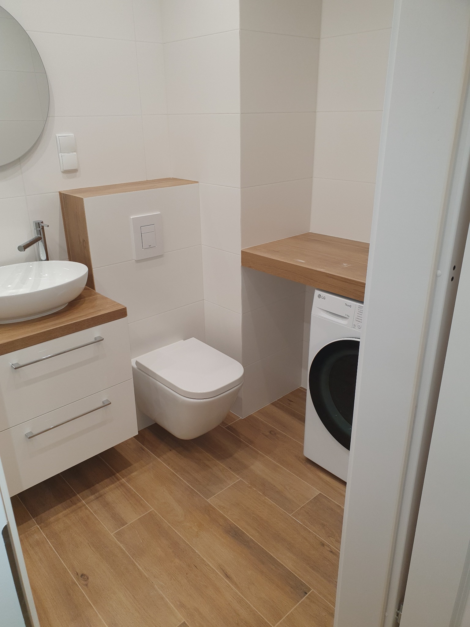 Nowoczesna łazienka w jasnych kolorach z drewnianymi akcentami: umywalka nablatowa, podwieszana toaleta, pralka pod blatem z drewna, brązowe płytki podłogowe.