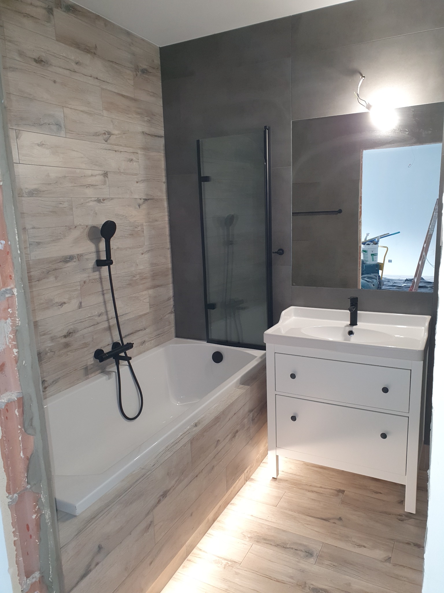 Ukończona łazienka z wanną, czarną armaturą i białą szafką umywalkową. Widoczne płytki imitujące drewno na ścianach i podłodze, oraz szara ściana z lustrem. Przez lustro widać fragment rusztowania.